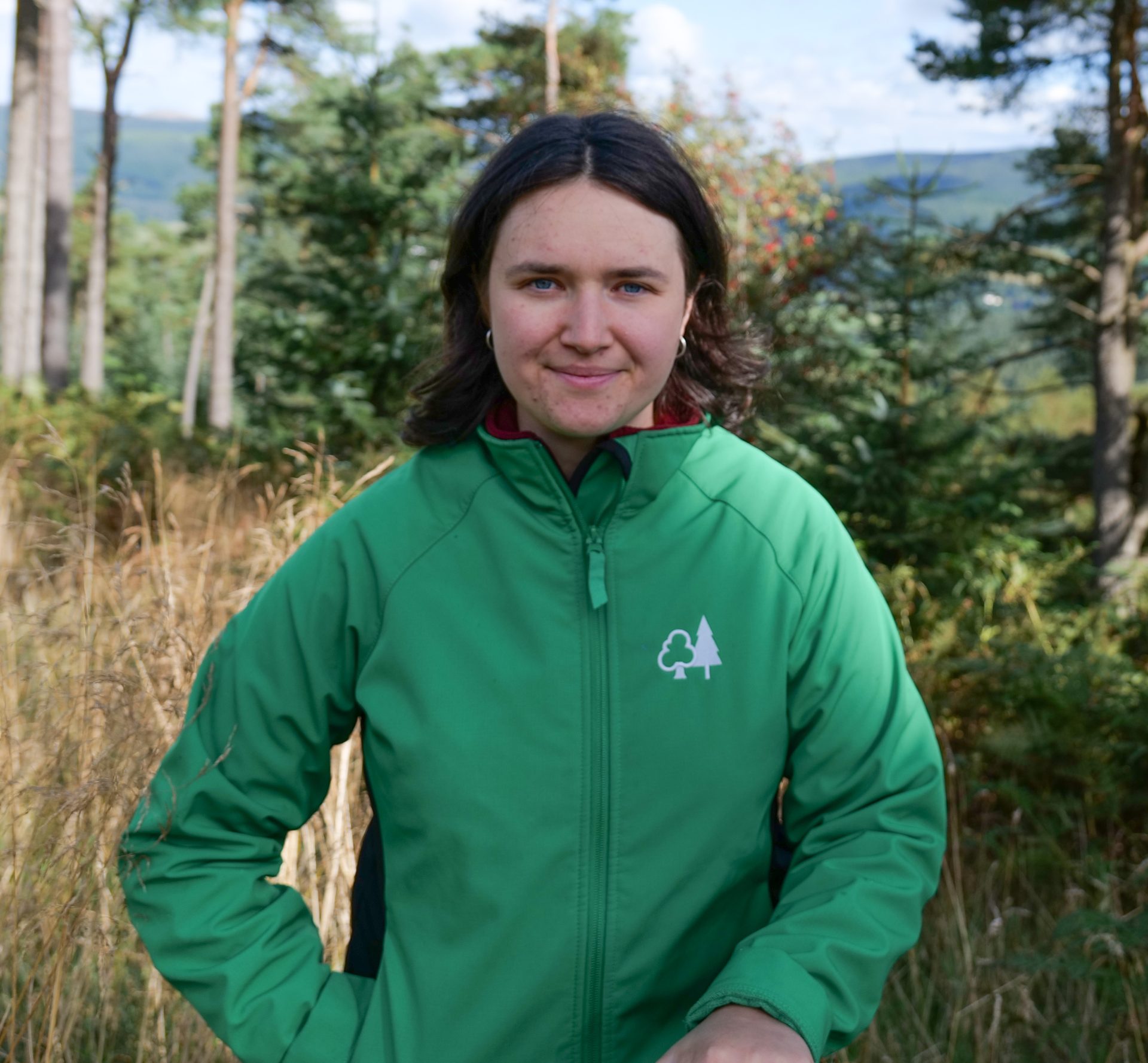 Mirabelle Ostle profile picture, she wears a green Forest Research jacket with trees in the background.