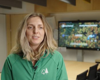 Suzanne Robinson, Climate Impacts Modeller, speaks to an interviewer, with a conference table and screen in the background with an aerial view of a forest.