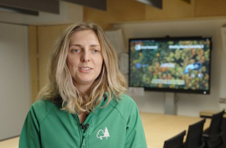 Suzanne Robinson, Climate Impacts Modeller, speaks to an interviewer, with a conference table and screen in the background with an aerial view of a forest.