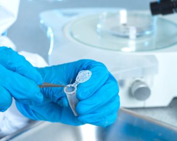 Two hands in blue gloves with scientific materials in the background. The hands are dealing with a sample in a small plastic tube.