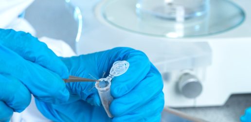 Two hands in blue gloves with scientific materials in the background. The hands are dealing with a sample in a small plastic tube.