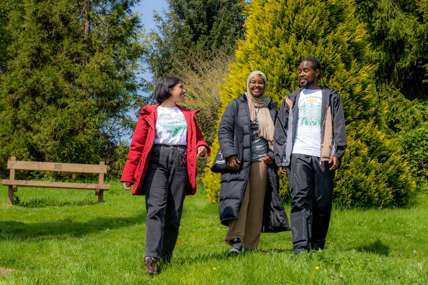 Three people in woods walk towards camera with smiles on their faces. One wears a red jacked and the other two wear black jackets. It's sunny and the scene is lush and green. 