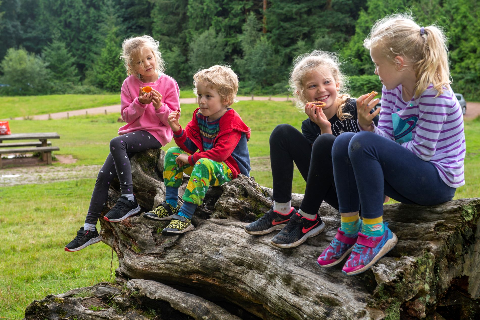 Four children in colourful clothing sit on a log with woodlands in tha background. They are eating a snack and smiling.