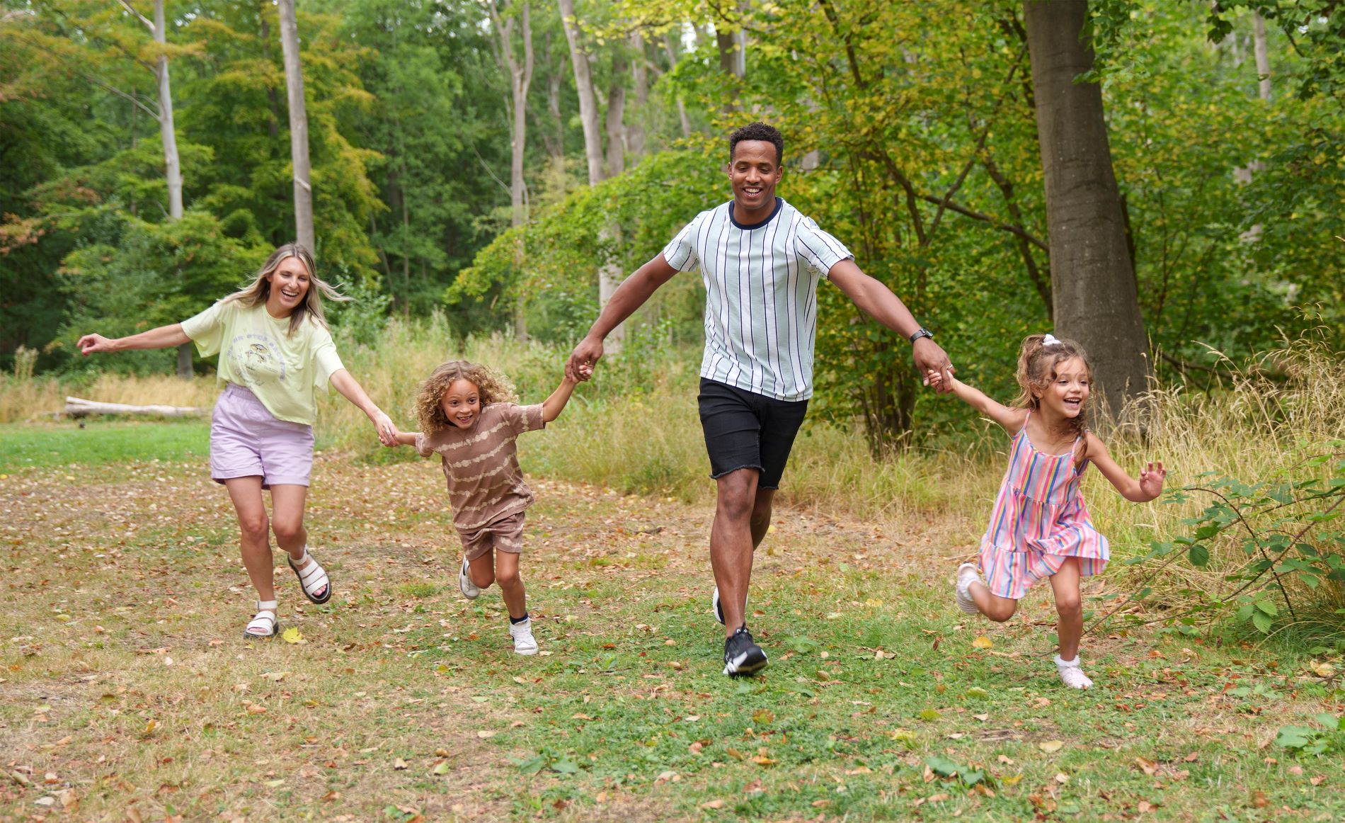 A mother and father with their two daughters run hand in hand in a woodland. They are smiling and laughing. It is autumnal with leaves on the ground and trees turning yellow. 