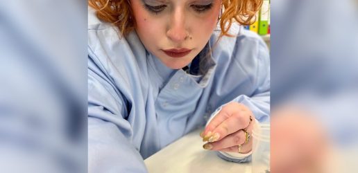 Danae is in a blue lab coat and is inspecting an insect on a white surface.