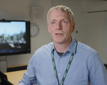 Forest Research's Bruce Nicol speaking to camera with a screen on in the background showing the damaging impact of wind on forests.