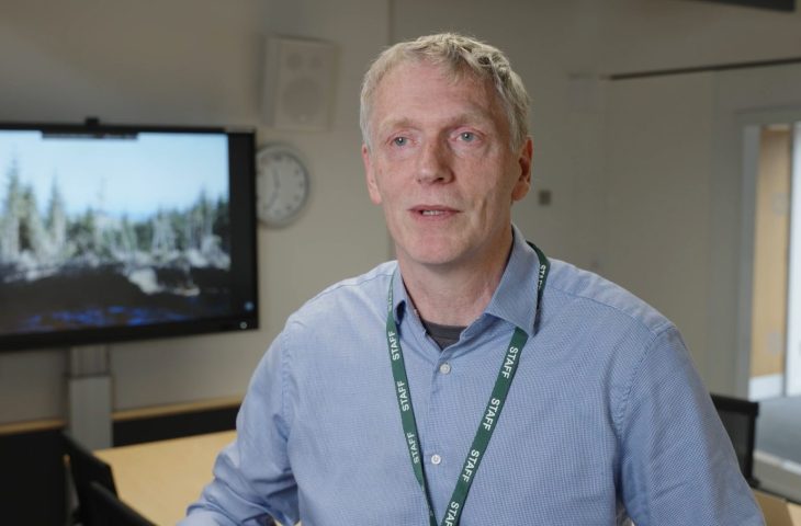 Forest Research's Bruce Nicol speaking to camera with a screen on in the background showing the damaging impact of wind on forests.