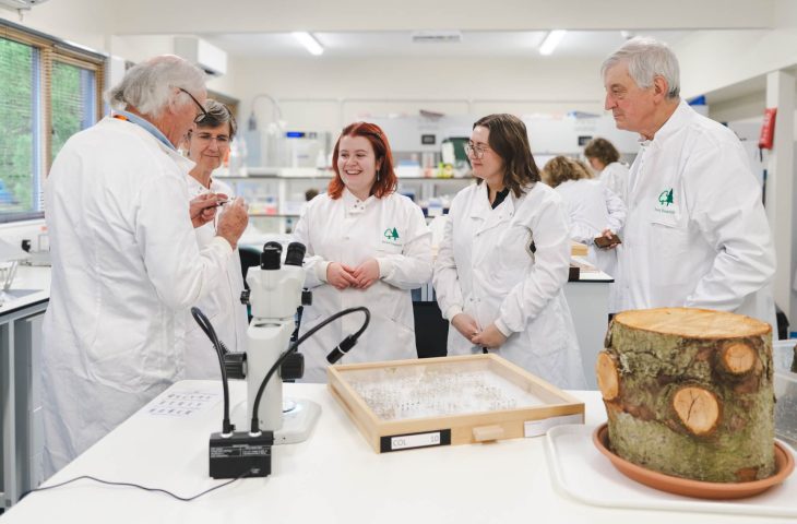 Volunteers gather round chatting to lab technician about samples. Microscope, pinned insect case and log in foreground.