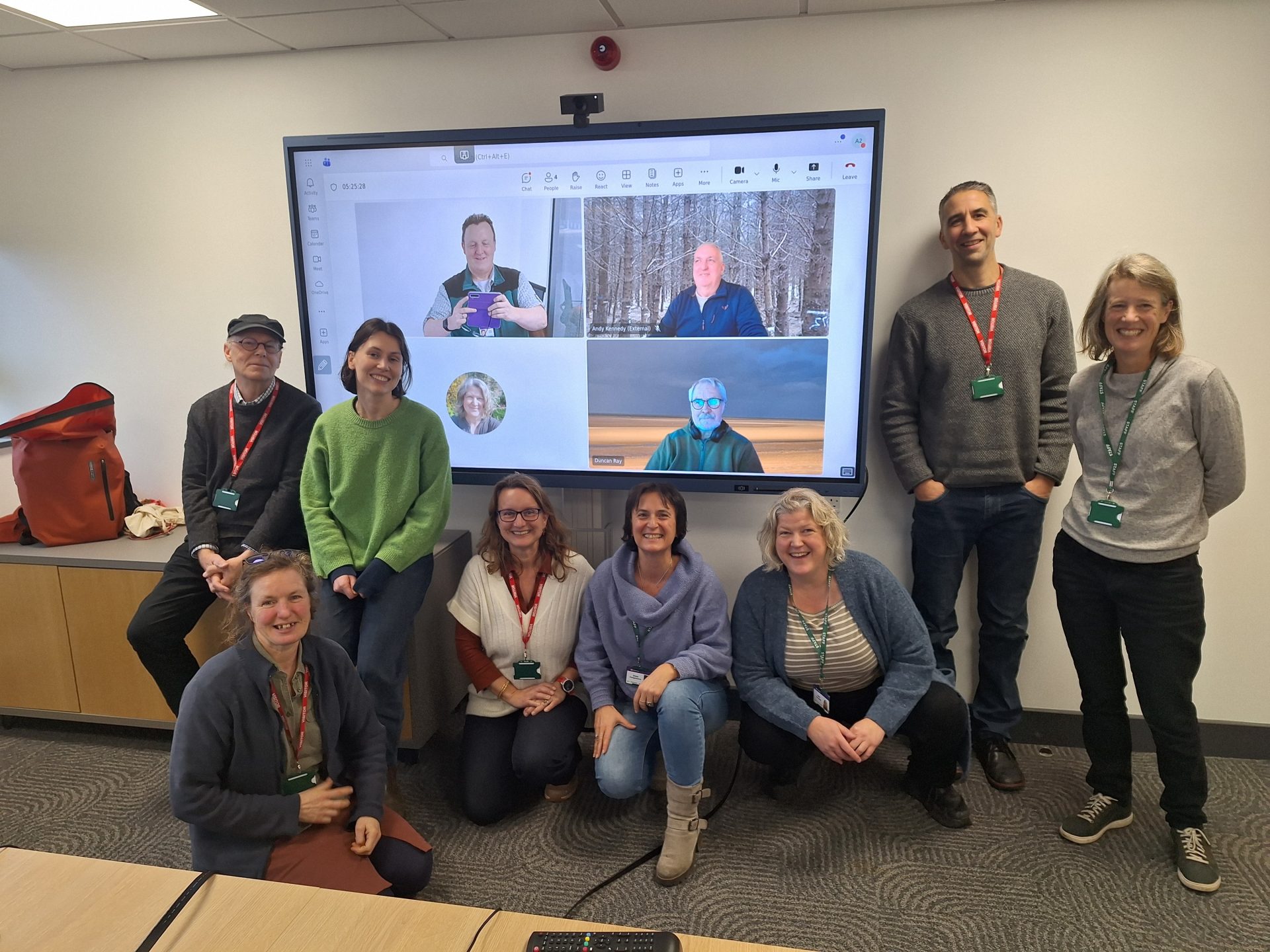 A group of people pose for a photo around a screen which has people on it. 